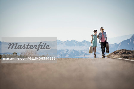 A young couple, man and woman, on a tarmac road in the desert carrying cases.