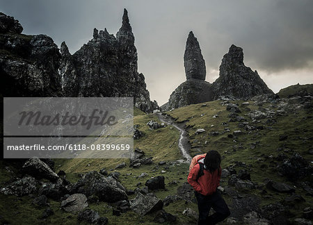 Two people with rucksacks on a narrow path rising to a dramatic landscape of rock pinnacles on the skyline towering above them, under an overcast sky with low cloud.