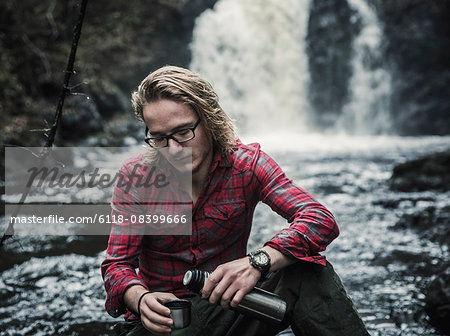 A young man sitting pouring a hot drink from a flask by a fast flowing stream.