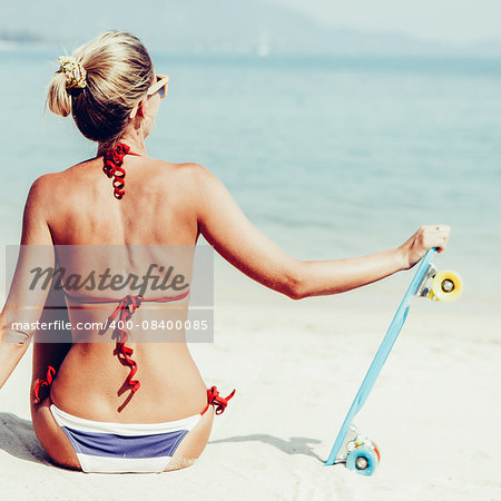 Suntanned lady in blue and white striped bikini sits on sandy beach and holding her penny board