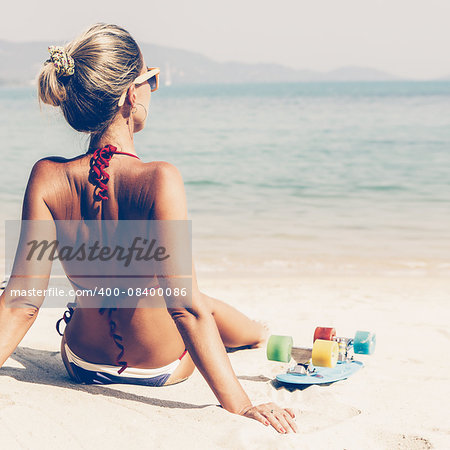 Beautiful sporty girl in blue and white striped bikini sits on sandy beach and sunbathing near her penny board