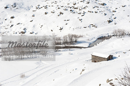 Remote stone mountain hut on an alpine slope next to river covered in snow