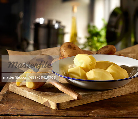 Potatoes being peeled with a knife with the peeled ones on a plate on a wooden board in a kitchen