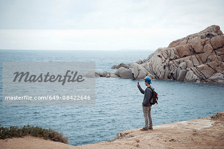 Young man standing on cliff using smartphone to photograph ocean, Costa Smeralda, Sardinia, Italy