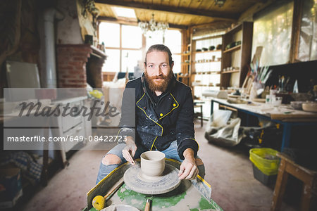 Bearded mid adult man in workshop sitting at pottery wheel making clay pot, looking at camera smiling