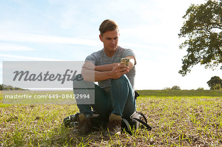 Young man sitting on backpack reading smartphone texts in field