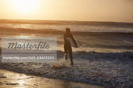 Young male surfer carrying surfboard running into sea, Devon, England, UK