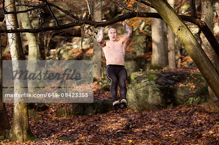 Full length front view of tattooed mid adult man in forest doing pull up on tree