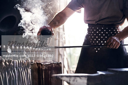 Blacksmith pouring hot liquid over wrought iron in forge