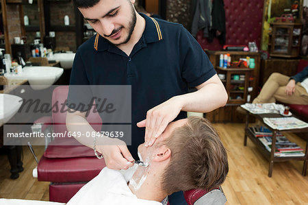 Young man in barbershop looking down shaving customer with straight razor