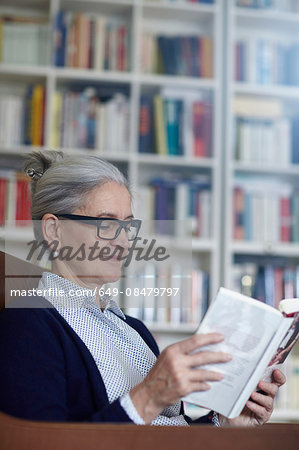 Grey haired mature woman reading book from bookshelves