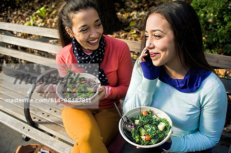 Young adult female twins sitting on park bench chatting on smartphone and eating lunch