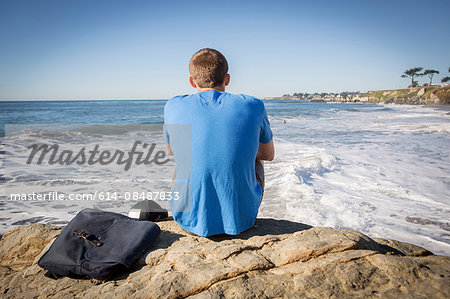 Young man sitting by rock, looking out to sea, rear view