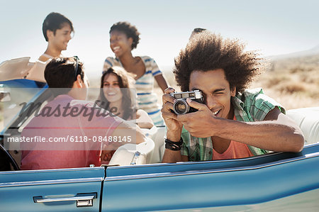 A group of friends in a pale blue convertible on the open road, one holding a camera and taking photographs.