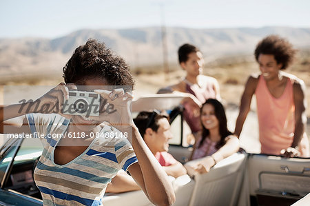 A group of friends by a pale blue convertible on the open road, on a flat plain surrounded by mountains, one holding a camera.