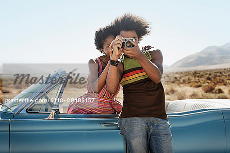 A young couple, man and woman by a pale blue convertible on the open road, holding a camera.