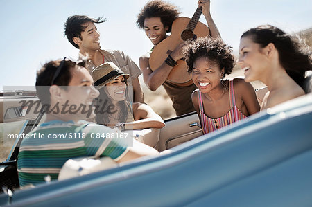 A group of friends in a pale blue convertible on the open road, driving across a dry flat plain surrounded by mountains.