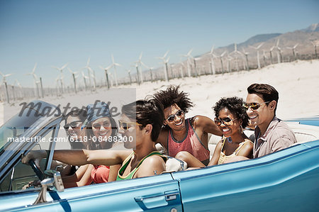 A group of friends in a pale blue convertible on the open road, driving across a dry flat plain surrounded by mountains.