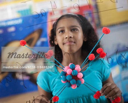 A girl holding a molecular structure and looking at a board of equations and formulae in the classroom.
