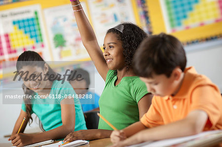 A group of young girls and boys in a classroom, classmates. A girl raising her hand.