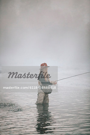 A woman fisherman fly fishing, standing in waders in thigh deep water.