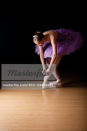 Young female ballerina wearing a lilac colored tut adjusting her shoes against black background