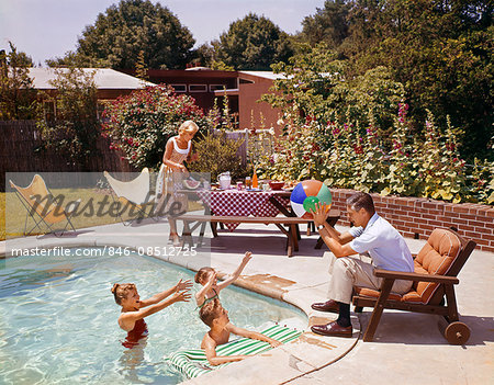 1950s 1960s MAN FATHER TOSSING BALL TO THREE CHILDREN IN SWIMMING POOL MOTHER FIXING PICNIC LUNCH SUBURBAN BACKYARD PATIO