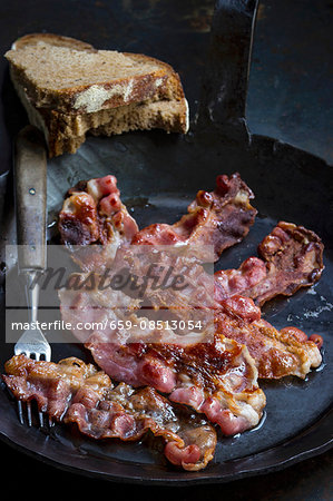 Fried bacon in a cast iron pan with a fork and slices of bread