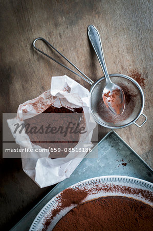 Chocolate cake on a metal tray with utensils for sprinkling cocoa powder on a wooden table