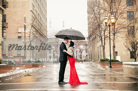 A woman in a long red evening dress with fishtail skirt and a fur stole, and a man in a suit, kissing under an umbrella in the city.