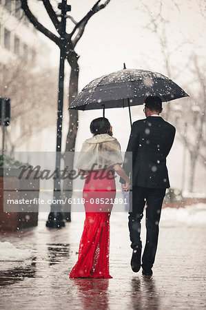 A woman in a long red evening dress with fishtail skirt and a fur stole, and a man in a suit, walking through snow in the city.