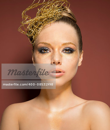 beauty shoot of very pretty woman with glossy golden creative make-up and accessory in the hairdo. She is covering her nude breast and looking in camera