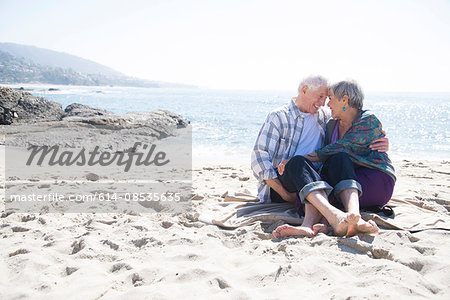 Senior couple sitting on beach, hugging, face to face, smiling