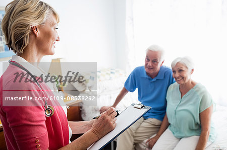 Nurse writing on clipboard and senior couple sitting on a bed