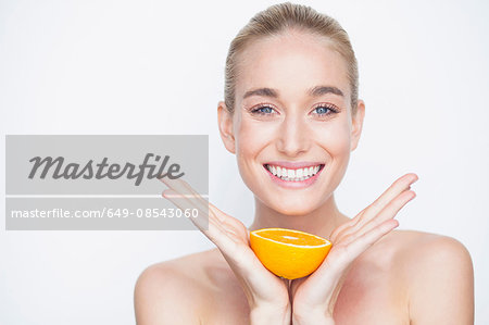 Bare shouldered woman holding half an orange looking at camera smiling