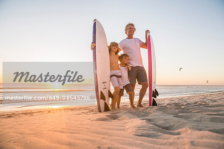 Full length view of father and sons standing on beach holding surfboard at sunset