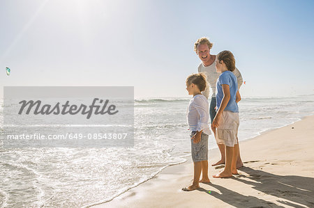 Full length side view of father and sons on beach looking away at ocean Full length side view of father and sons on beach looking away at ocean