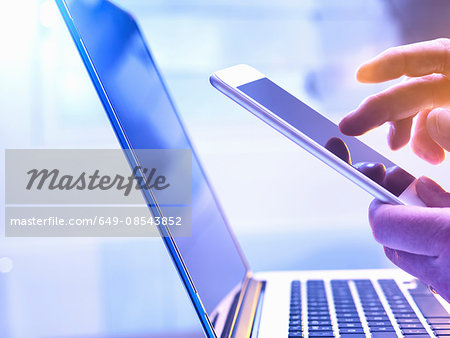 Man operating a touch screen mobile phone with a laptop in the background.