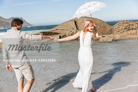 Couple holding hands, walking on coastline holding umbrella looking over shoulder smiling
