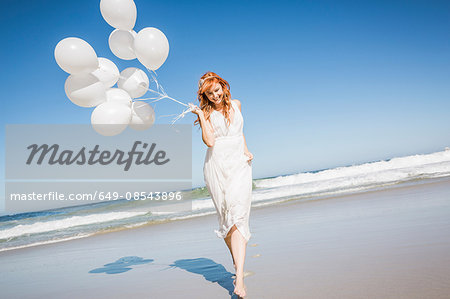 Full length front view of woman on beach wearing white dress holding balloons