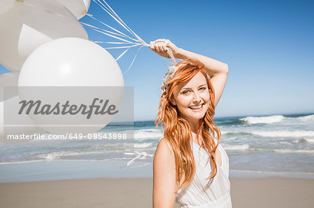 Red haired woman on beach holding balloons looking at camera smiling