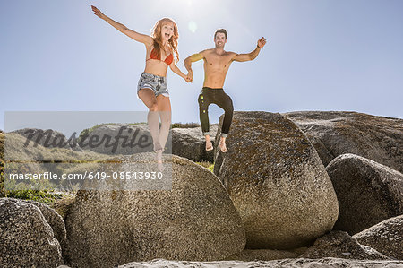 Couple outdoors, holding hands, jumping off of rock onto sand