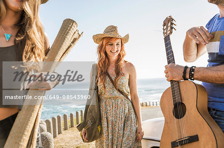 Three mid adult friends carrying acoustic guitar and mat on coast road, Cape Town, South Africa