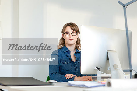 Young woman wearing eyeglasses sitting at desk using computer looking away smiling