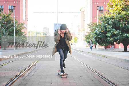 Young male skateboarder adjusting earphones whilst skateboarding on tramway