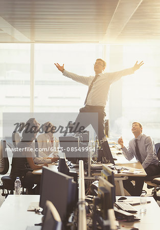 Colleagues watching exuberant businessman celebrating on top of desks in office