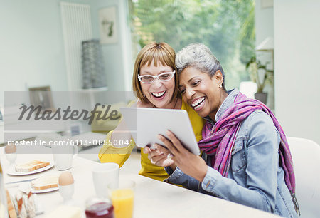 Laughing mature women sharing digital tablet at breakfast table