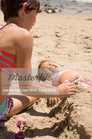 Girl burying other girl in sand on beach