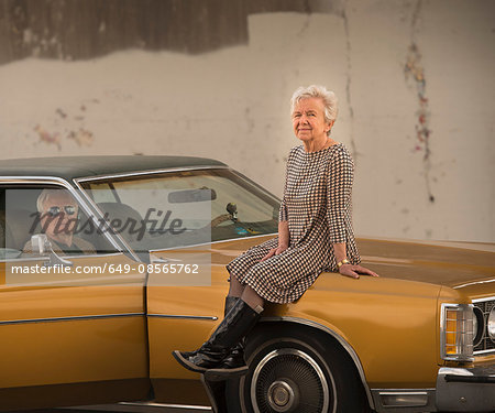 Portrait of cool senior woman sitting on bonnet of husbands classic automobile