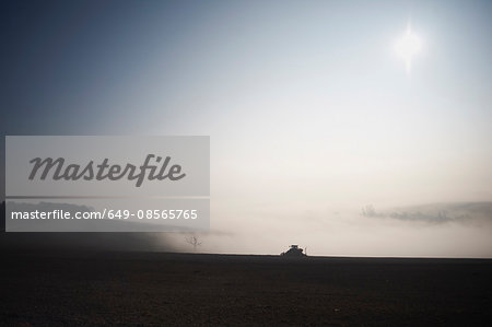 Silhouetted tractor ploughing field in morning mist